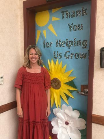 Picture of Angie Killian in front of her office door decorated with words saying,  "Thank you for helping us grow."  The door is covered with paper with a yellow sun and red, yellow, and white paper flowers. 