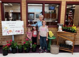 pictured is the owner of Olsen Garden Shoppe with a sign, flowers, and garden display for the kids to enjoy
